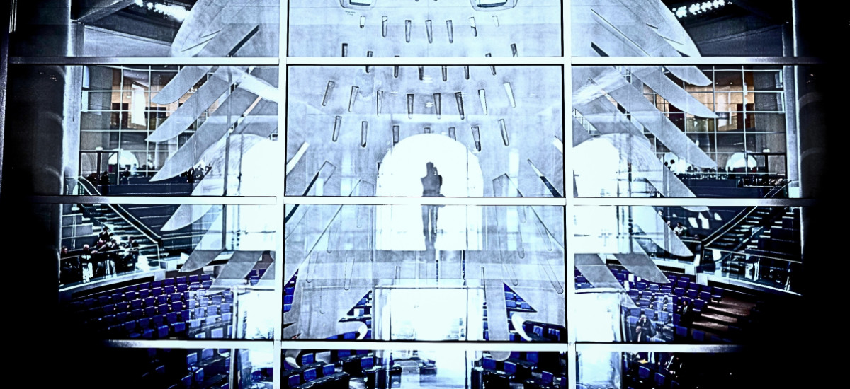 Blick durch die Glasfassade in den Plenarsaal des Deutschen Bundestags. Im Vordergrund sind die großen, stilisierten Bundesadler-Elemente auf der Glaswand zu sehen. Dahinter erkennt man die blauen Sitzreihen des Plenarsaals, Treppen und einzelne Personen. Im Zentrum steht silhouettenhaft eine Person vor einem hell erleuchteten Durchgang. Das Bild ist kontrastreich und in kühlen Blau- und Grautönen gehalten.