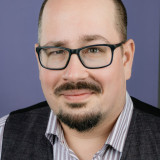 Thomas Ponier-Kröhl Ein Bild von Thomas Ponier-Kröhl vor einem lila Hintergrund. Er trägt ein gestreiftes Hemd und eine Weste.