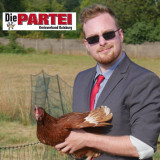 Ein Mann mit Sonnenbrille und einem Huhn in seinen Händen, welcher auf einem Feld steht. In der oberen linken Ecke steht ein Schrift zu mit Die PARTEI Kreisverband Duisburg und einer Grafik der Lebensretter-Statue