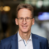 Ein schlanker, sportlich wirkender, Mann mit brauner Kurzhaarfrisur, Brille, weißem Hemd und blauem Jacket