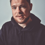 Profilbild von Moritz Riedacher. Es zeigt ihn sitzend, mit geraden Blick in die Richtung des/der Betrachter:in blickend. Er trägt einen schwarzen Kapuzenpullover, hat einen Bart und kurze Haare. 