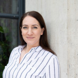 Eva Miriam Fuchs Eva Miriam Fuchs steht vor einer Betonwand und Fensterwand und blickt lächelnd gerade in die Kamera