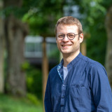 Konstantin Lotz Der Kandidat Konstantin Lotz im Porträt. Er trägt ein blaues Hemd, hat blonde Haare und trägt eine Brille. Im Hintergrund sind Bäume von einem Park zu sehen.