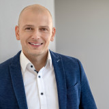Porträtfoto von Florian Bellaire im dunkelblauen Sakko und weißem Hemd vor neutralem Hintergrund