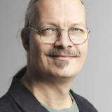 Frederic Müller Der Kandidat auf dem Foto trägt Brille und Oberlippenbart, hat die Haare zu einem sogenannten Pferdeschwanz gebunden. Er trägt unter einem schwarzen Jacket einen grünen Pullover.