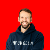 Auf dem Bild sieht man Philipp Dehne vor einem glatten roten Hintergrund. Man sieht ihn vom Kopf bis etwa zum Bauch, er blickt frontal in die Kamera. Philipp trägt einen dunkelblauen Hoodie, auf dem in weißen Buchstaben NEUKÖLLN steht.