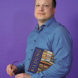 Hendrik Schäfer steht vor violettem Hintergrund und hält ein Buch in der Hand.