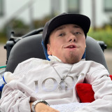 Samuel Niklas Scholz Das Bild zeigt eine Person in einem Rollstuhl. Sie trägt ein helles Sweatshirt mit der Aufschrift „JOOP“, eine silberne Kette und eine schwarze Kappe. Die Person hat einen Beatmungsschlauch, der seitlich befestigt ist. Im Hintergrund sind unscharf Gebäude und eine Treppe zu erkennen. Ein rotes Kissen und eine weiche Decke sind ebenfalls sichtbar.