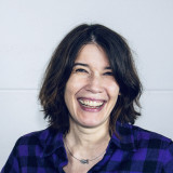 Katja Bühring Katja Bühring schaut offen und mit einem Lachen den Betrachter an. Sie trägt ein lila-schwarz kariertes Karohemd und eine Kette mit einem kleinen Fisch als Anhänger.