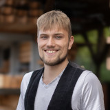 Lars Biesenthal zusehen in seiner Zimmerkluft, vor einem Holzlager. 