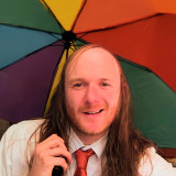 Paul Friedrich Ulrich Heitmann Das Bild zeigt einen jungen Mann mit langen Haaren und Halbglatze. Er trägt ein weißes Hemd mit einer schlecht gebundenen roten DIE PARTEI-Krawatte. Er lächelt etwas verpeilt und hat einen Regenbogenschirm in der Hand.