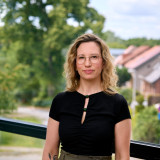 Es ist eine Frau mit blonden Haaren und Brille auf dem Foto zu sehen. Sie trägt ein schwarzen T-Shirt und eine dunkelgrüne Hose. Im Hintergrund sind verschwommen Böume und Hausdächer zu erkennen. 