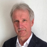 Das ist ein Foto von Jürgen Verkennis. Ich habe blaue Augen. Einen kurzen Oberlippenbart. Mittelscheitel und die Haare gehen bis knapp über die Ohren. Altersbedingt habe ich echte graue Haare. Ich trage gerade ein weißes Hemd und ein blaugraues Jackett. Im Hintergrund sieht man meine weiße Wohnzimmertapete.