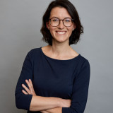 Lisett mit verschränkten Armen. Sie trägt ein dunkelblaues Shirt mit Dreiviertel-Ärmeln und lächelt herzlich.