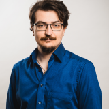 Edgar Krez Porträt eines Mannes mit dunklen Haaren, Brille und einem gepflegten Bart. Er trägt ein königsblaues Hemd mit offenem Kragen und eine silberne Kette. Sein Gesichtsausdruck ist ruhig und selbstbewusst. Der Hintergrund ist neutral und hell.