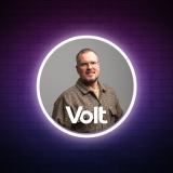 Kandidat Heiko trägt ein kariertes Hemd in Erdfarbtönen, um ihn herum ein weißer Kreis und die Aufschrift Volt. Der Hintergrund ist lila und beinhaltet eine Brandmauer.