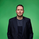 Marcel Richter Das Bild zeigt Marcel Richter in einem dunklen Anzug mit einem dunkelgrauen Shirt darunter. Er hat kurze, dunkelblonde Haare und eine freundliche, entspannte Gesichtsausdruck. Der Hintergrund ist ein einfarbiger, kräftiger grüner Farbton. Das Porträt ist professionell aufgenommen, mit guter Beleuchtung und einem klaren Fokus auf das Gesicht der Person.