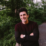 Das Bild zeigt Simon Schmitz lächelnd an ein Holzbalkengeländer auf einem Wanderweg gelehnt. Er hat die Arme vor der Brust verschränkt und im Hintergrund ist ein schöner grüner Baum und grüne Sträucher zu sehen.