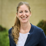 Sylvia Ganz im blauen Blazer und weißem Shirt, die langen Haare zum Zopf gebunden. Sie lächelt freundlich direkt in die Kamera. Im Hintergrund eine weich verschwommene Parkszene mit dichtem Gebüsch und einer ockerfarbenen Mauer.