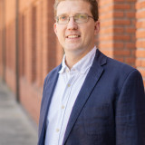 Thomas Völker Das Bild zeigt einen jungen Mann Mitte Dreißig mit Brille und welligem braunen Haar. Er lächelt und trägt ein weißes Hemd mit blauem Sakko darüber. Er steht vor einer roten Ziegelsteinmauer.