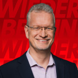 Weißer Mann mit Brille und grau-meliertem Haar. Im dunklen Sakko mit fliederfarbenem Hemd. Lächelt vor einem roten Hintergrund in die Kamera. 