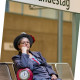 Andrea Schulteisz sitzt auf einer Bank im U-Bahnhof Bundestag. Neben ihr steht eine Tasche mit einer großen Uhr vorne drauf. Die Zeiger stehen auf kurz vor 12. 