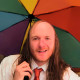 Das Bild zeigt einen jungen Mann mit langen Haaren und Halbglatze. Er trägt ein weißes Hemd mit einer schlecht gebundenen roten DIE PARTEI-Krawatte. Er lächelt etwas verpeilt und hat einen Regenbogenschirm in der Hand.