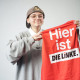 "Loo" Elena Krein Bild von Kandidatin "Loo" Elena Krein mit roter Weste in der Hand auf der "Hier ist DIE LINKE" steht