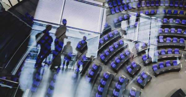 Ein Bild des Bundestag-Plenarsaals, schemenhaft sind Menschen in einer Spiegelung erkennbar.